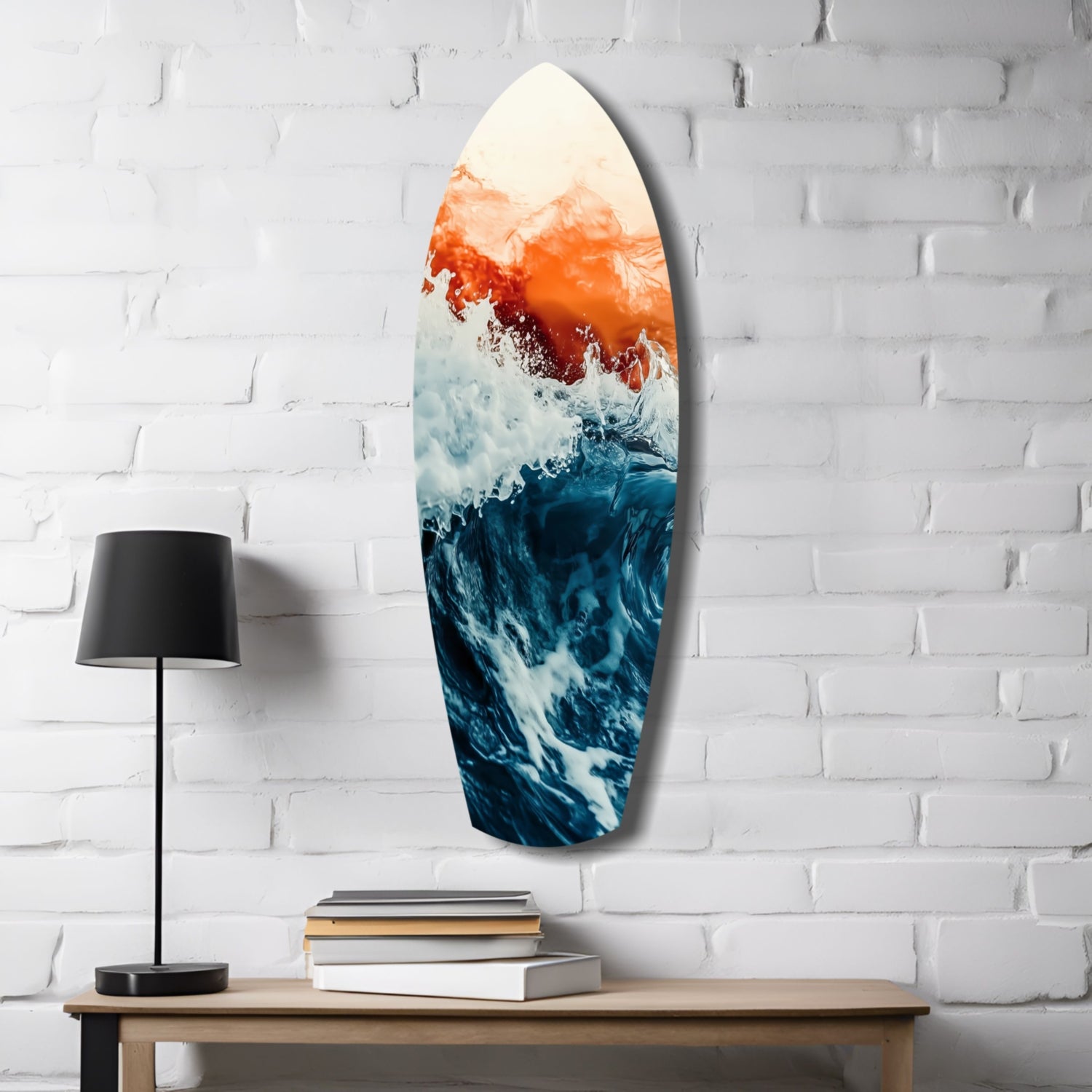 Surfboard-shaped glass wall art displaying ocean waves in deep blue and white with orange-red sunset gradient, hung on white brick beside wooden table with lamp and books.