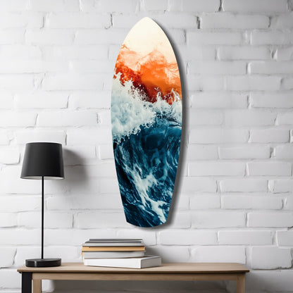 Surfboard-shaped glass wall art displaying ocean waves in deep blue and white with orange-red sunset gradient, hung on white brick beside wooden table with lamp and books.
