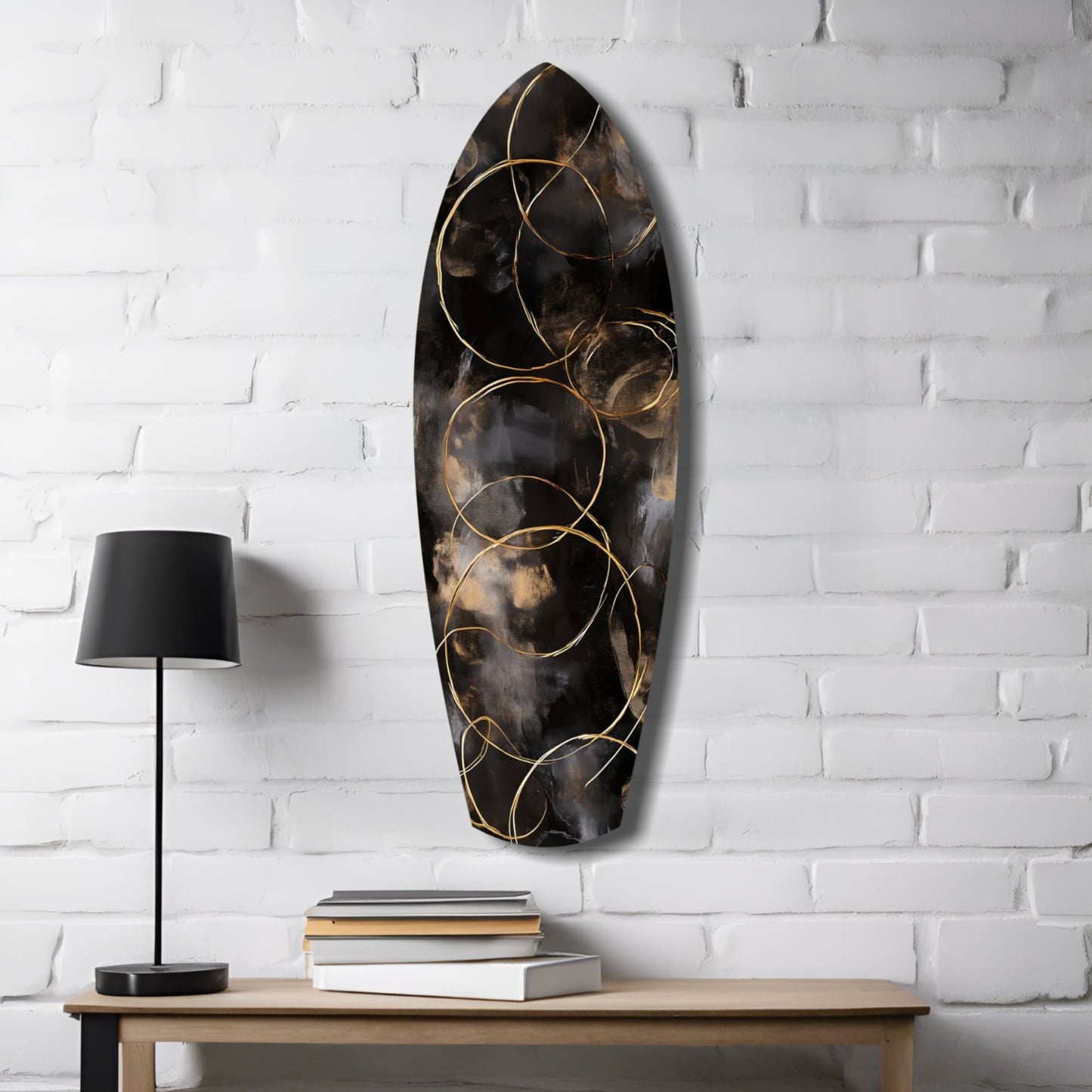Black and gold glass wall art with circular design displayed above wooden table with lamp and stacked books against white brick wall.