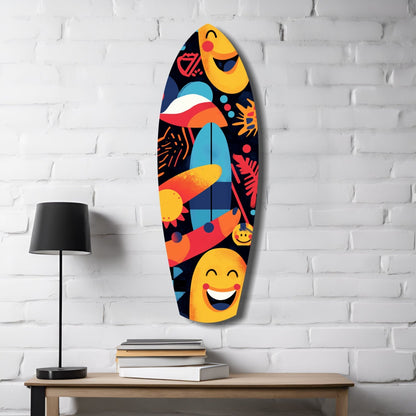 Colorful surfboard-shaped glass wall art with yellow smiling faces, geometric patterns, and vibrant orange, red, and blue designs displayed on white brick wall above wooden desk with lamp and books.