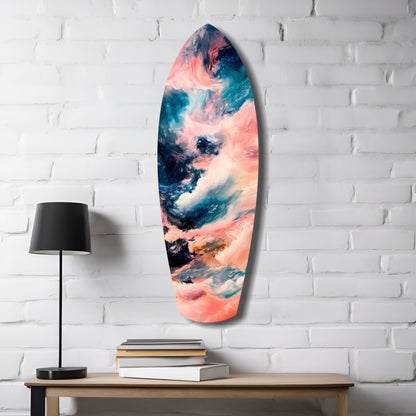 Surfboard-shaped glass wall art with coral, teal, and white abstract cloudy patterns displayed above a wooden desk with lamp and books.