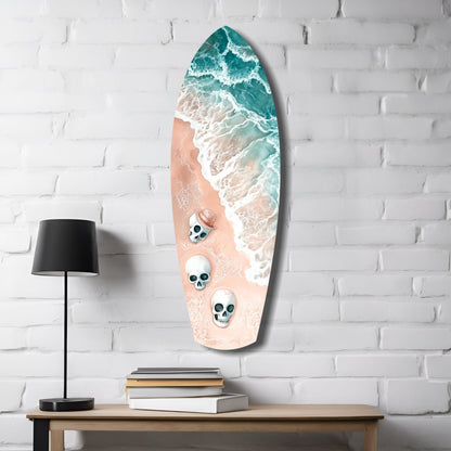 Surfboard-shaped glass wall art displaying aerial beach scene with turquoise ocean waves, sandy beach, and three skull designs, mounted on white brick wall beside lamp and books.
