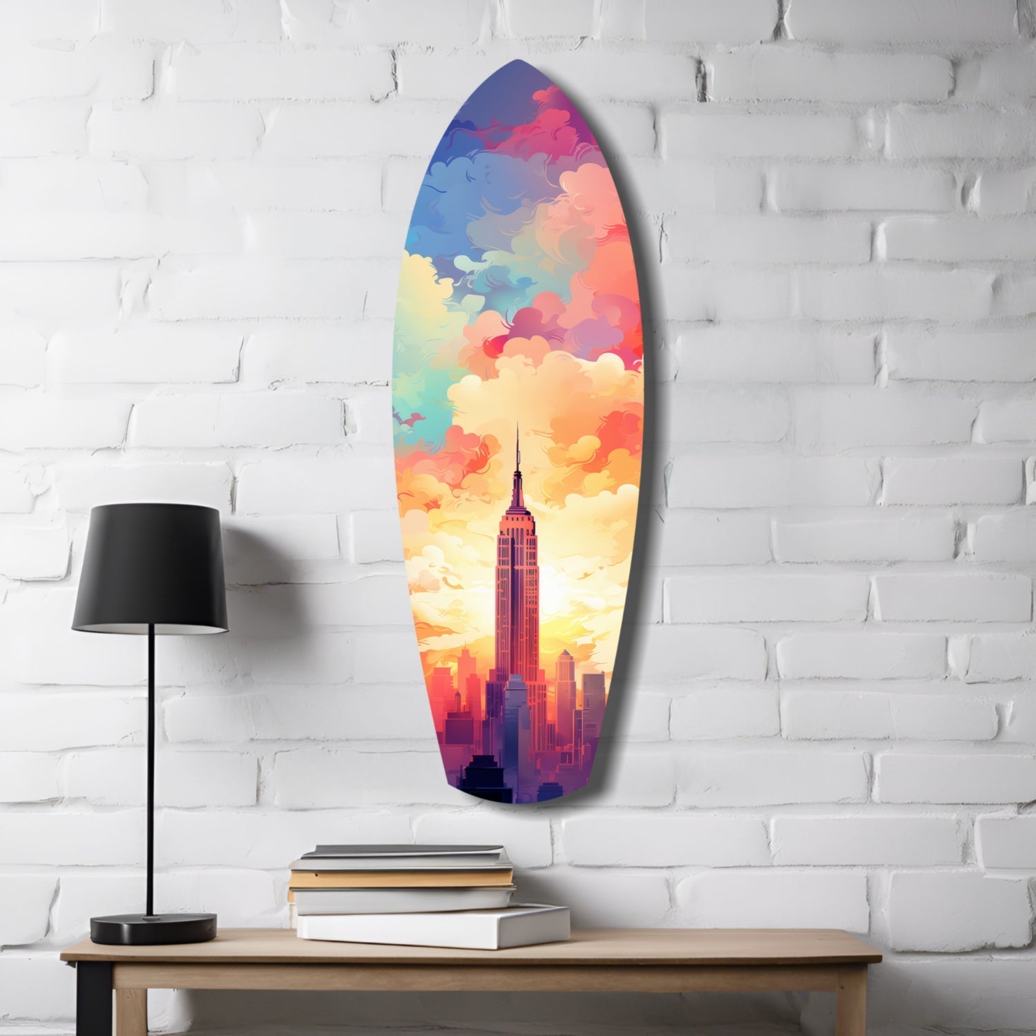 Surfboard-shaped glass wall art featuring Empire State Building silhouette against vibrant rainbow clouds, mounted on white brick wall above wooden desk with lamp and books.