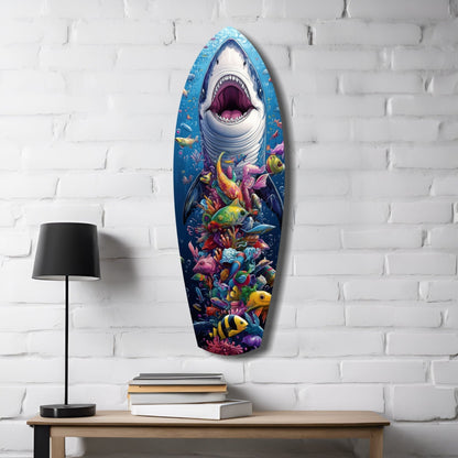 Surfboard-shaped glass wall art mounted on white brick displays a great white shark with open mouth above a colorful coral reef ecosystem, displayed above a wooden desk with lamp and books.