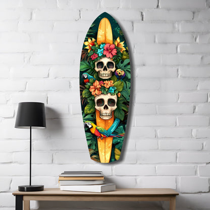 Surfboard-shaped glass art hanging on white brick wall above wooden desk with lamp and books, featuring two skulls surrounded by colorful flowers and a parrot.