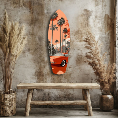 Orange surfboard-shaped glass wall art depicting a vintage red car and palm trees, displayed above a wooden bench against a textured concrete wall.