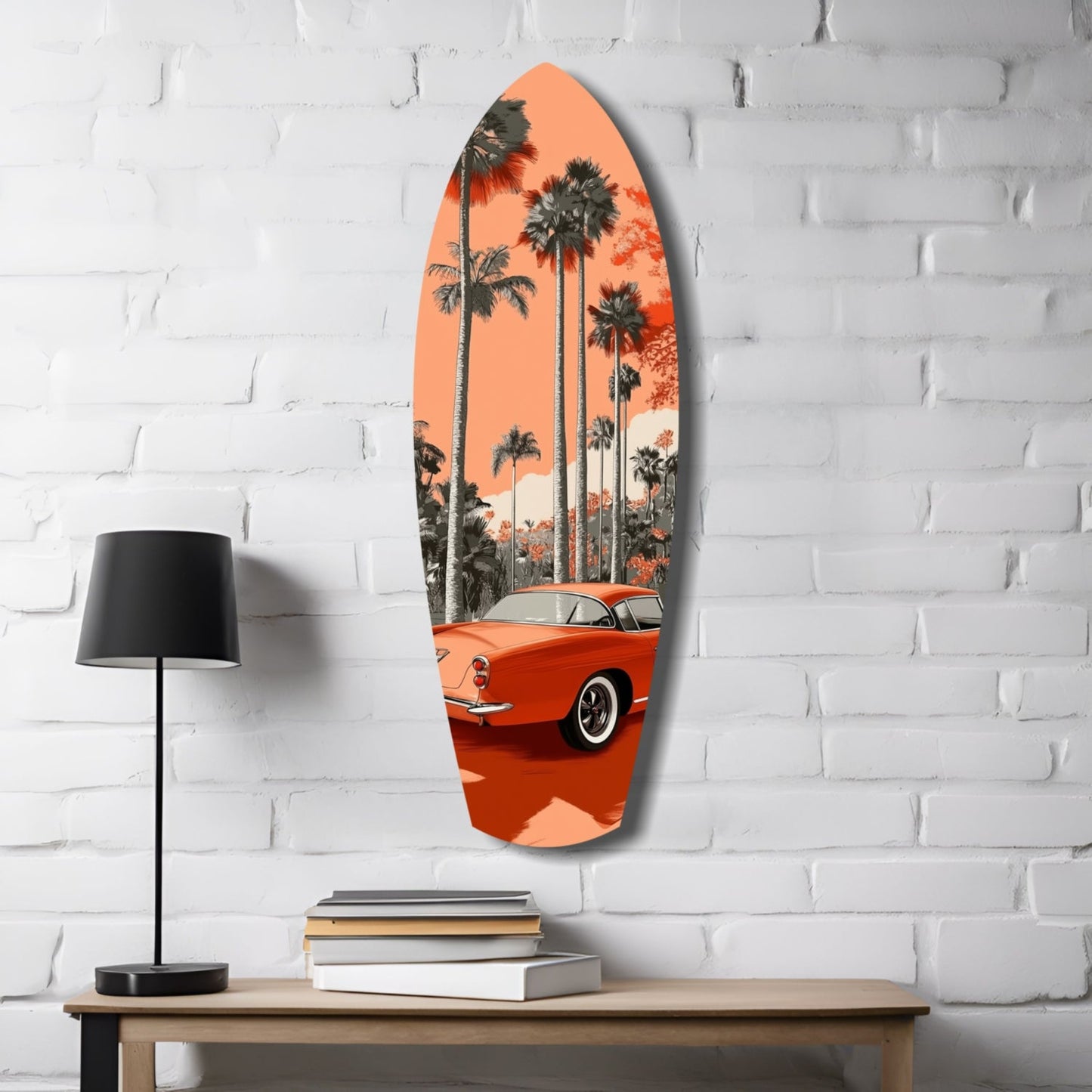Orange surfboard-shaped glass wall art with vintage red car and palm trees, displayed on white brick wall beside wooden table with lamp and books.
