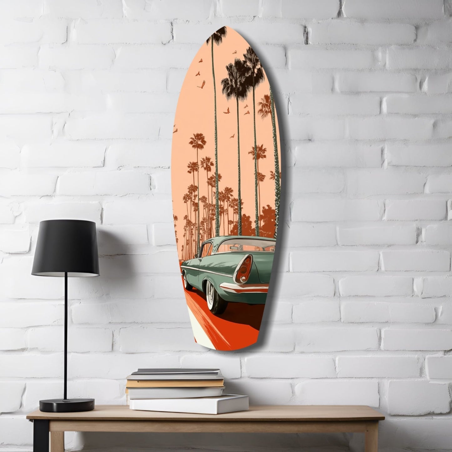 Surfboard-shaped glass wall art depicting a turquoise vintage car with palm trees against peachy sunset sky, displayed on white brick wall above wooden table with lamp and books.