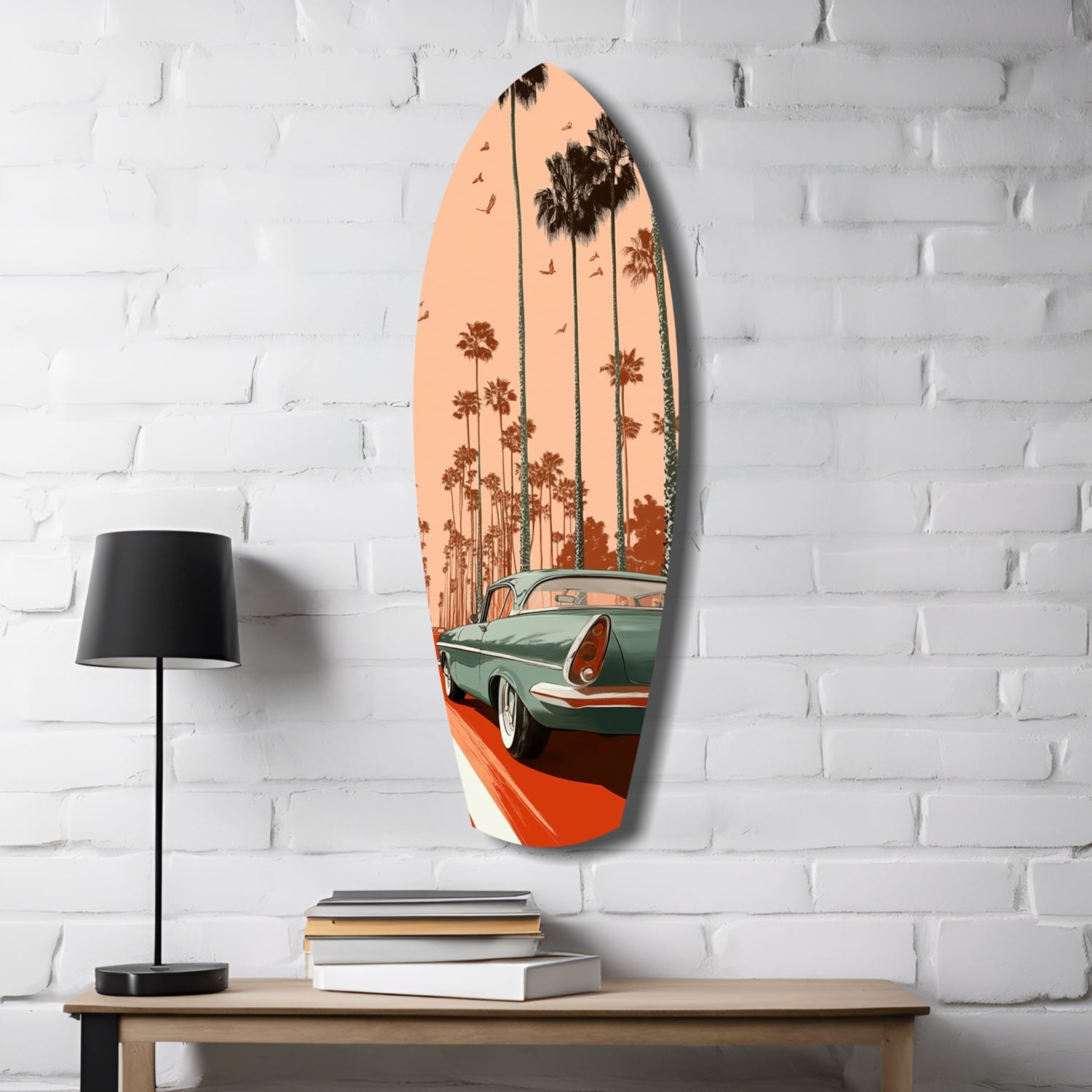 Surfboard-shaped glass wall art depicting a turquoise vintage car with palm trees against peachy sunset sky, displayed on white brick wall above wooden table with lamp and books.
