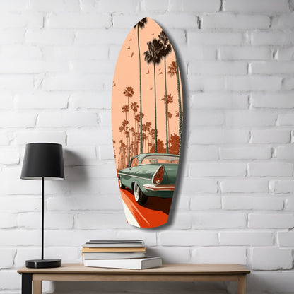 Surfboard-shaped glass wall art depicting a turquoise vintage car with palm trees against peachy sunset sky, displayed on white brick wall above wooden table with lamp and books.