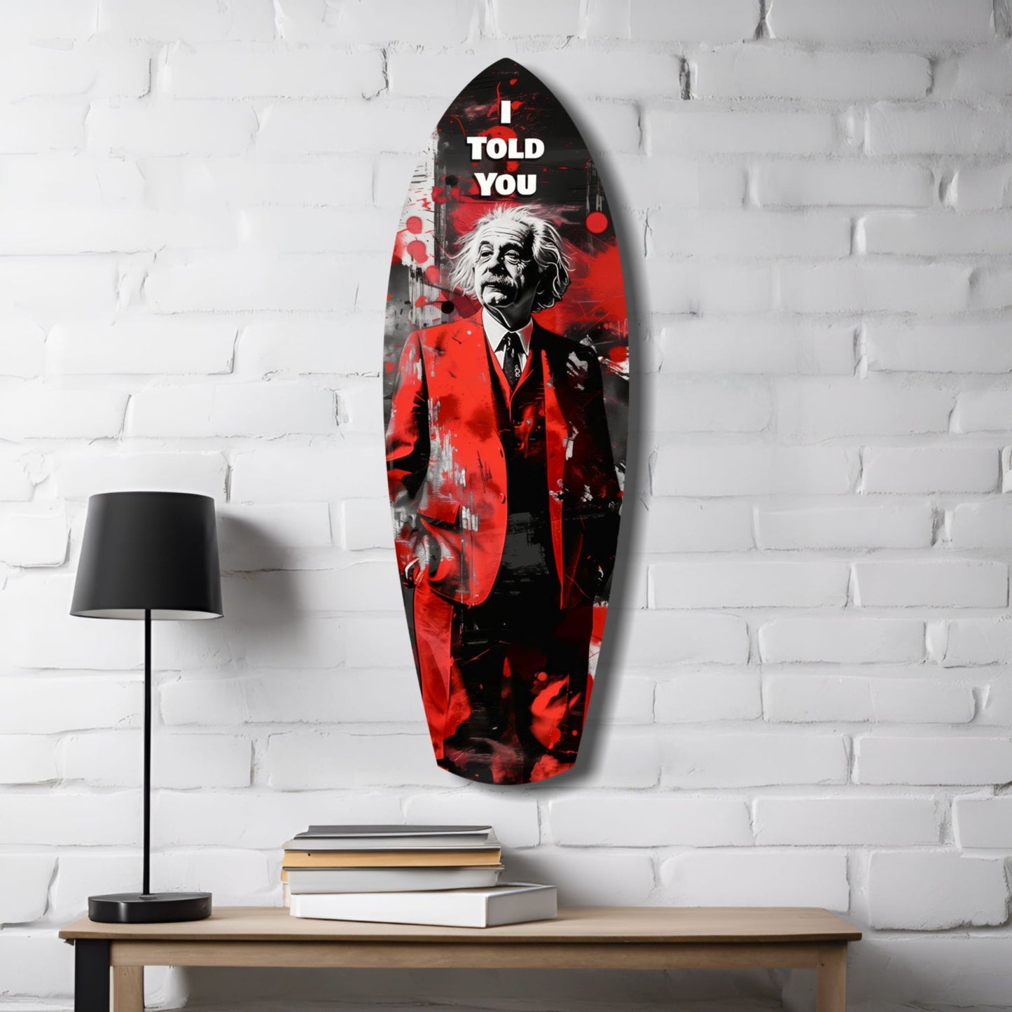 Red and black glass wall art depicting Einstein with "I TOLD YOU" text, displayed on white brick wall above wooden table with lamp and books.
