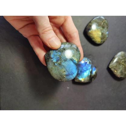 A hand holds a labradorite carved polished heart stone with iridescent blue and green flashes on a dark background