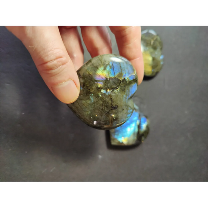 A hand holds a polished heart-shaped labradorite carved stone with iridescent blue and green flashes