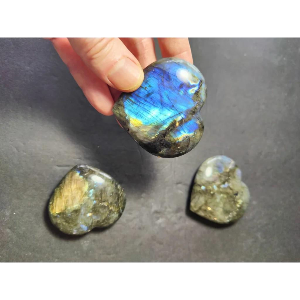 A hand holds a labradorite carved polished heart stone with blue and gold iridescent patterns and two stones nearby