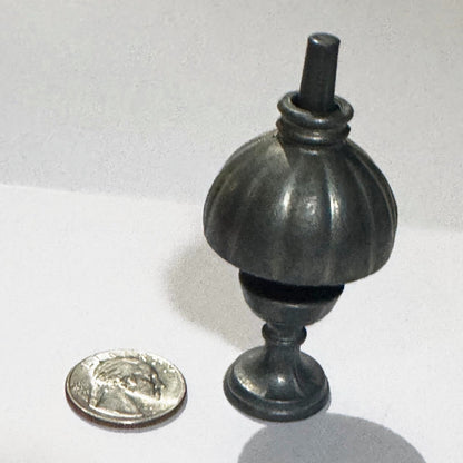 The miniature oil lamp from a higher angle, the tapered post rising through the ringed collar of its fluted shade, with a US quarter beside it for scale