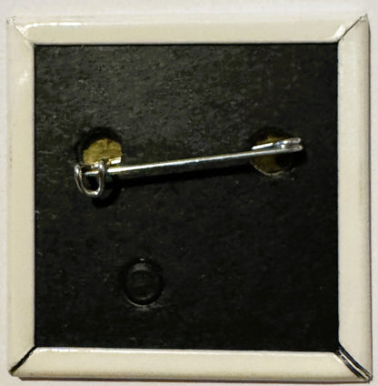 Silver-toned pin with brass circular elements mounted on black slate background in white frame.