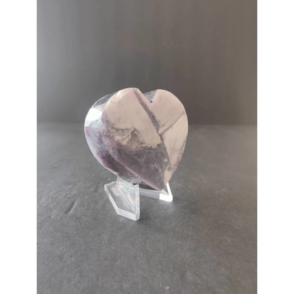 A translucent heart-shaped amethyst stone with purple and white patterns displayed on a clear acrylic stand from lepidolite tower heart sculptures