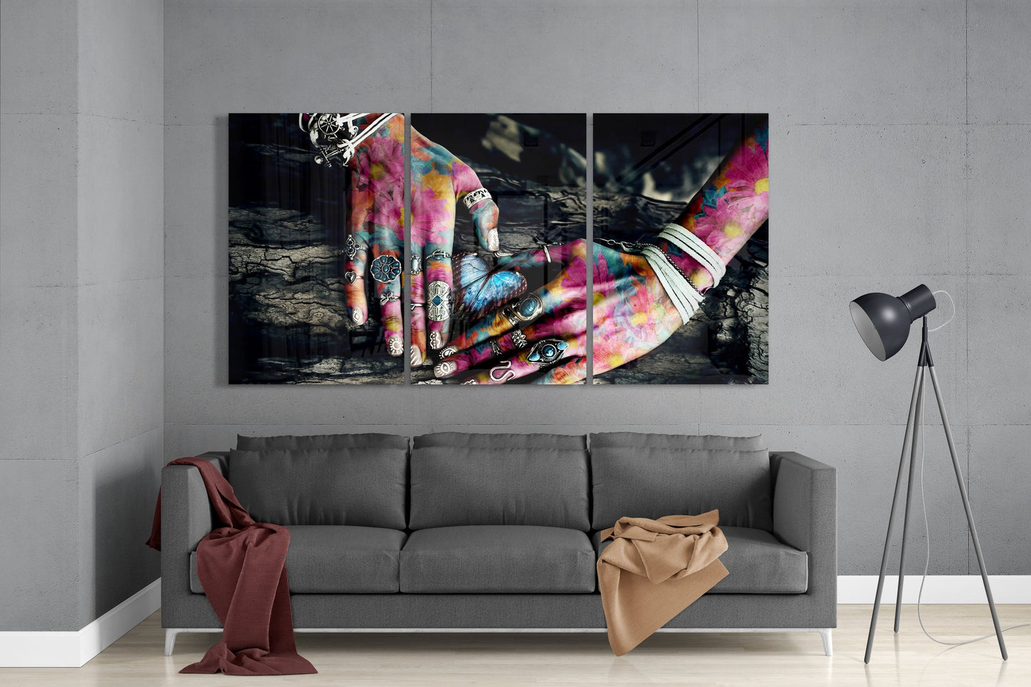 Triptych glass wall art depicting colorful tattooed hands with rings against dark textured background, displayed above gray sofa.