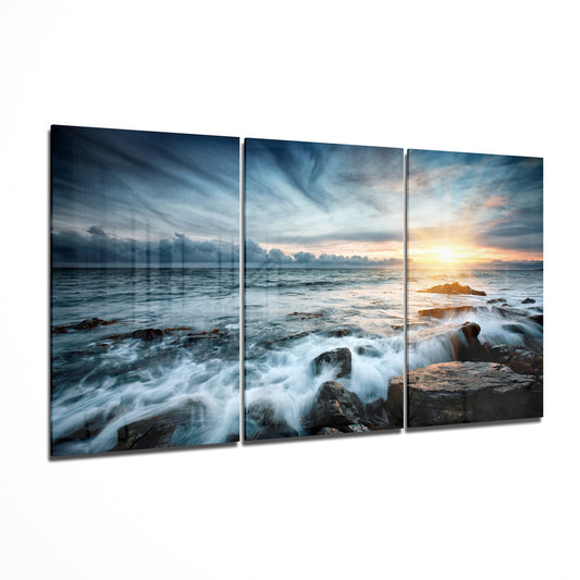 Three-panel glass wall art displaying a rocky coastline with crashing waves, dramatic cloudy sky, and golden sunset on the right panel.