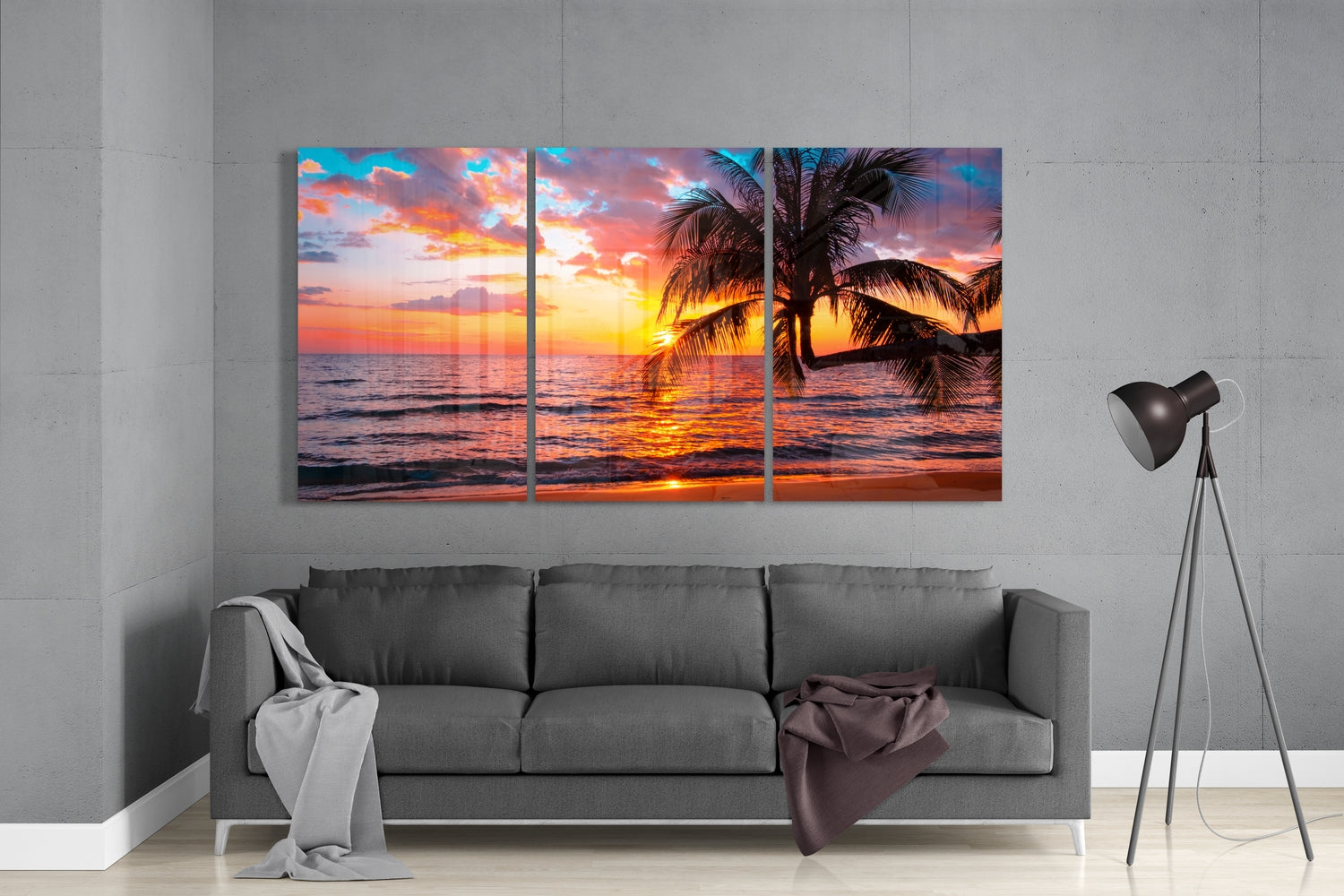 Three-panel glass wall art displaying a tropical sunset with palm trees and ocean, hung above a gray sofa in a modern living room.