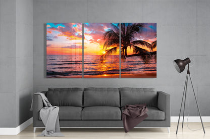 Three-panel glass wall art displaying a tropical sunset with palm trees and ocean, hung above a gray sofa in a modern living room.