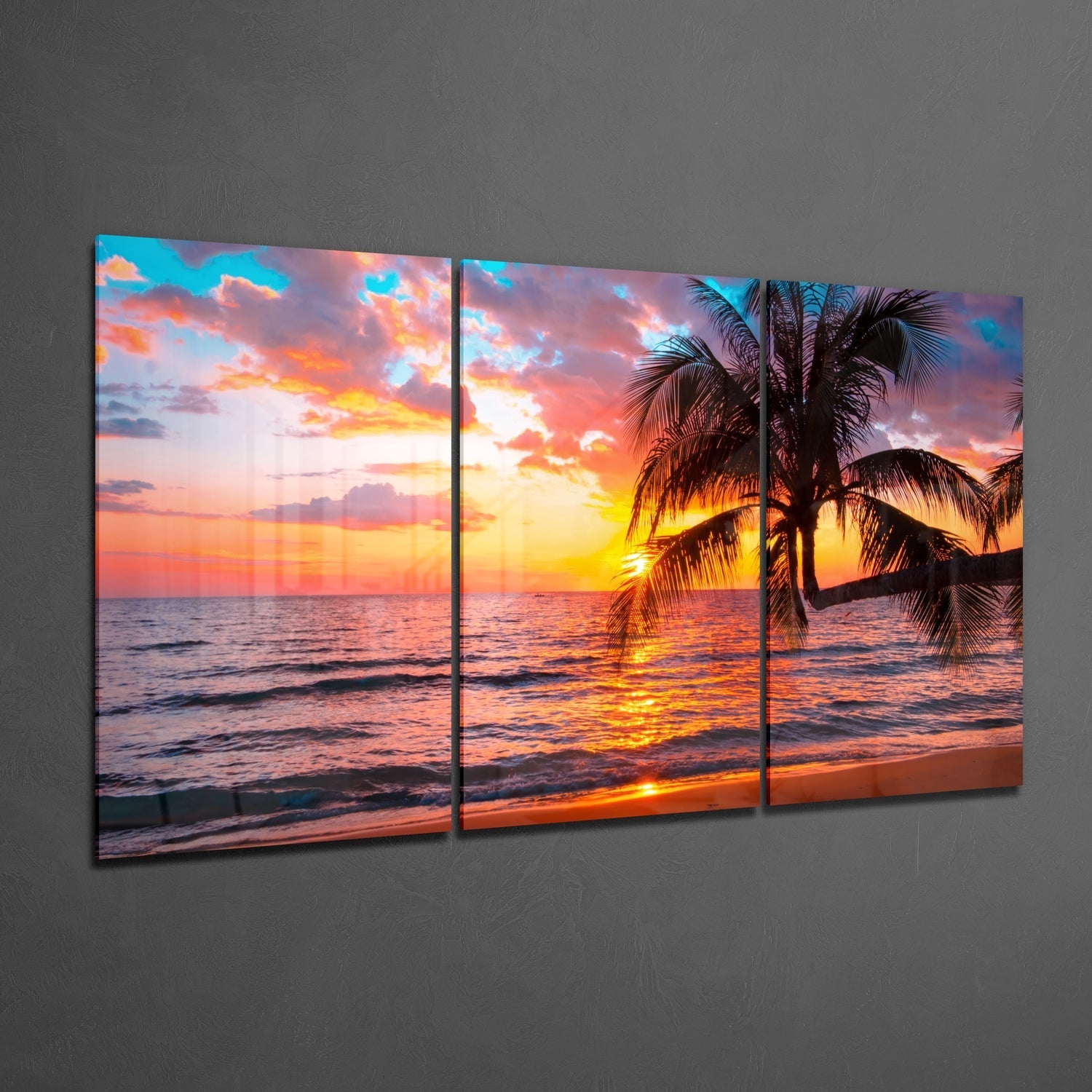 Three-panel glass wall art displaying a tropical beach sunset with vibrant orange and yellow sky, silhouetted palm trees, and ocean waves.