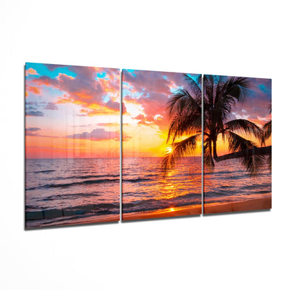Three-panel glass wall art displaying a tropical beach sunset with palm trees, golden sky, and ocean waves.
