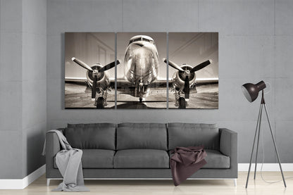 Three-panel sepia-toned glass artwork displaying front view of vintage twin-engine aircraft on tarmac, installed above gray sofa in modern interior.