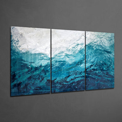 Three-panel glass wall art displaying turbulent water textures transitioning from white foam at top to deep teal and navy blue at bottom, mounted on gray wall.