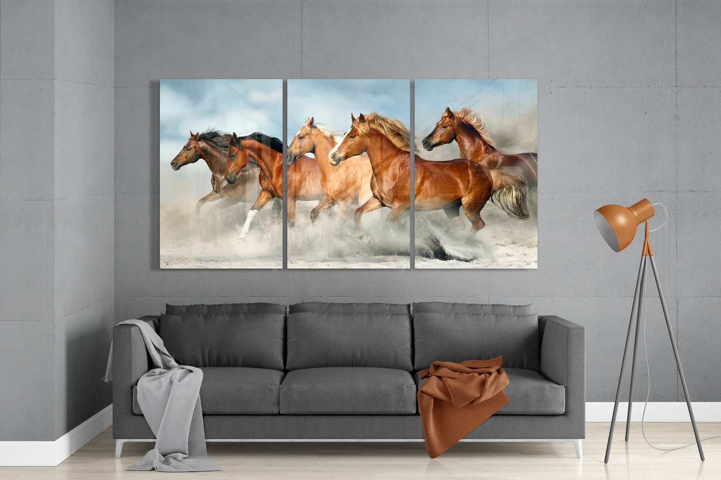 Three-panel glass wall art displaying brown horses galloping through dust, installed above gray sofa in modern living room.