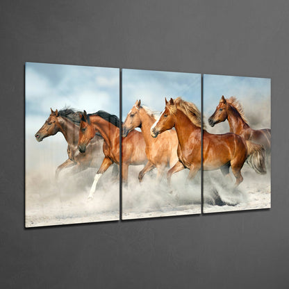 Three-panel glass wall art displaying brown horses galloping through dust against a cloudy sky backdrop, mounted on gray wall.
