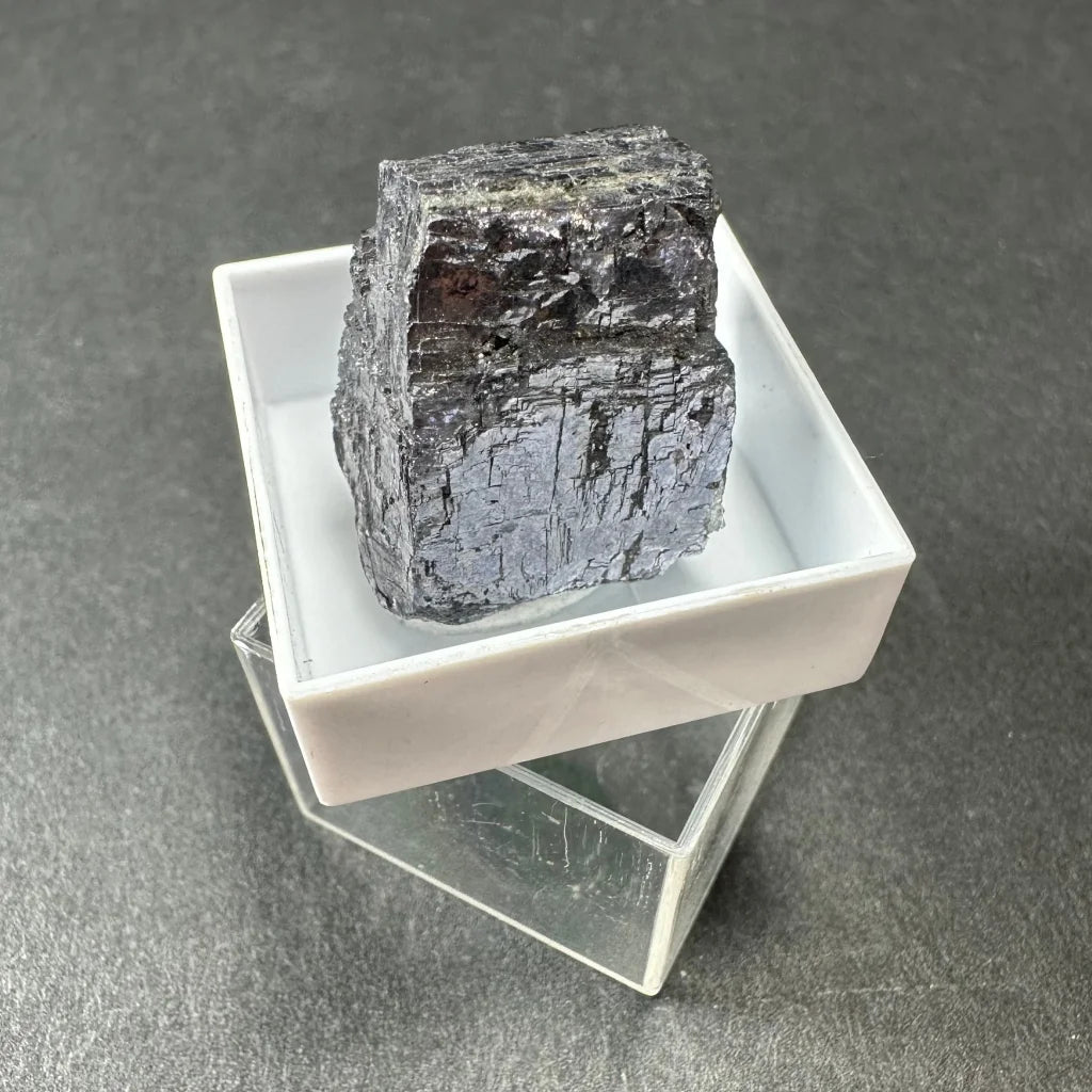 Dark Moroccan thumbnail specimen with metallic sheen in acrylic box