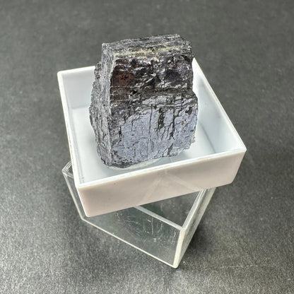 Dark Moroccan thumbnail specimen with metallic sheen in acrylic box
