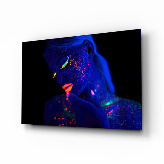 Woman's face in profile with neon blue, red, and yellow body paint and glitter against black background, displayed on glass wall art panel.