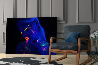 Neon portrait glass art displaying fluorescent blue, pink, and yellow face paint on a woman's face, displayed on a living room wall beside a wooden chair.
