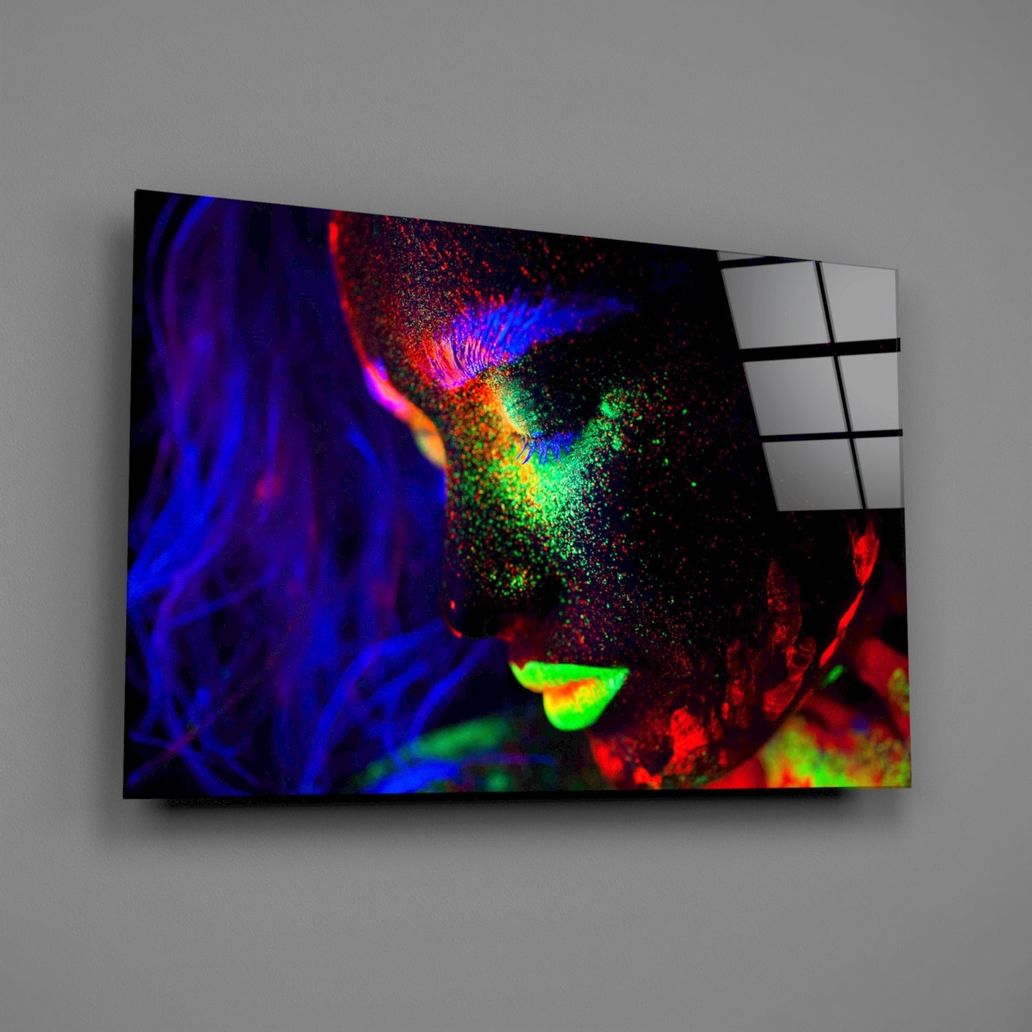 Angled view of colorful glass wall art panel displaying vibrant neon blue, red, and green abstract portrait with white grid panel visible on right edge.