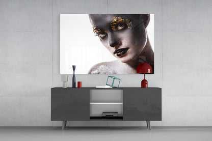 Glass wall art featuring a woman's face with gray skin, gold eyeshadow, and black lips displayed above a dark TV console.