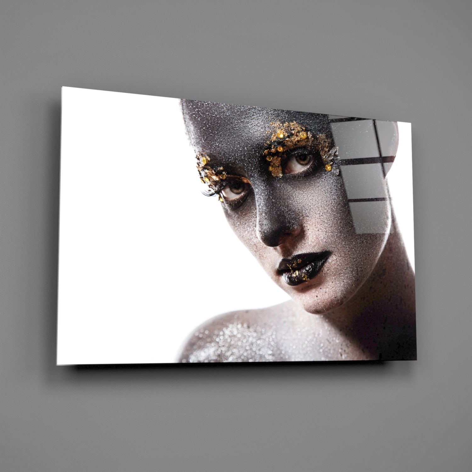 Glass wall art displaying a portrait of a woman's face with silver glitter makeup, gold accents around eyes, and black lips against white background.