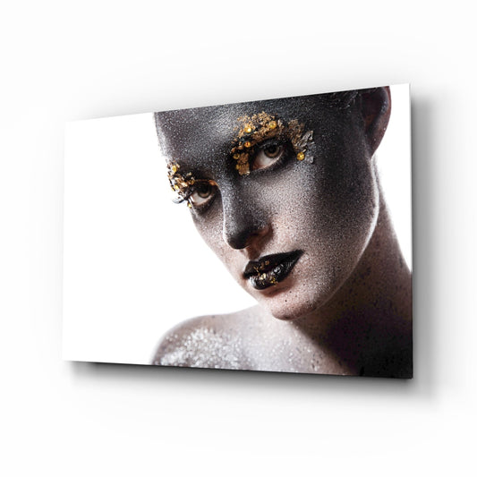 Woman's face with silver glitter makeup and gold leaf accents around eyes and lips, displayed on glass wall art.