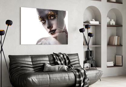 Glass wall art displaying close-up portrait of woman with gold eye makeup and white sparkles against white background, mounted above black leather sofa.