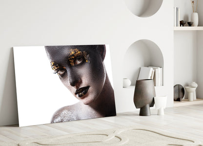 Glass wall art leaning against white wall displaying portrait of woman with gold leaf eyes and dark lips on white background.