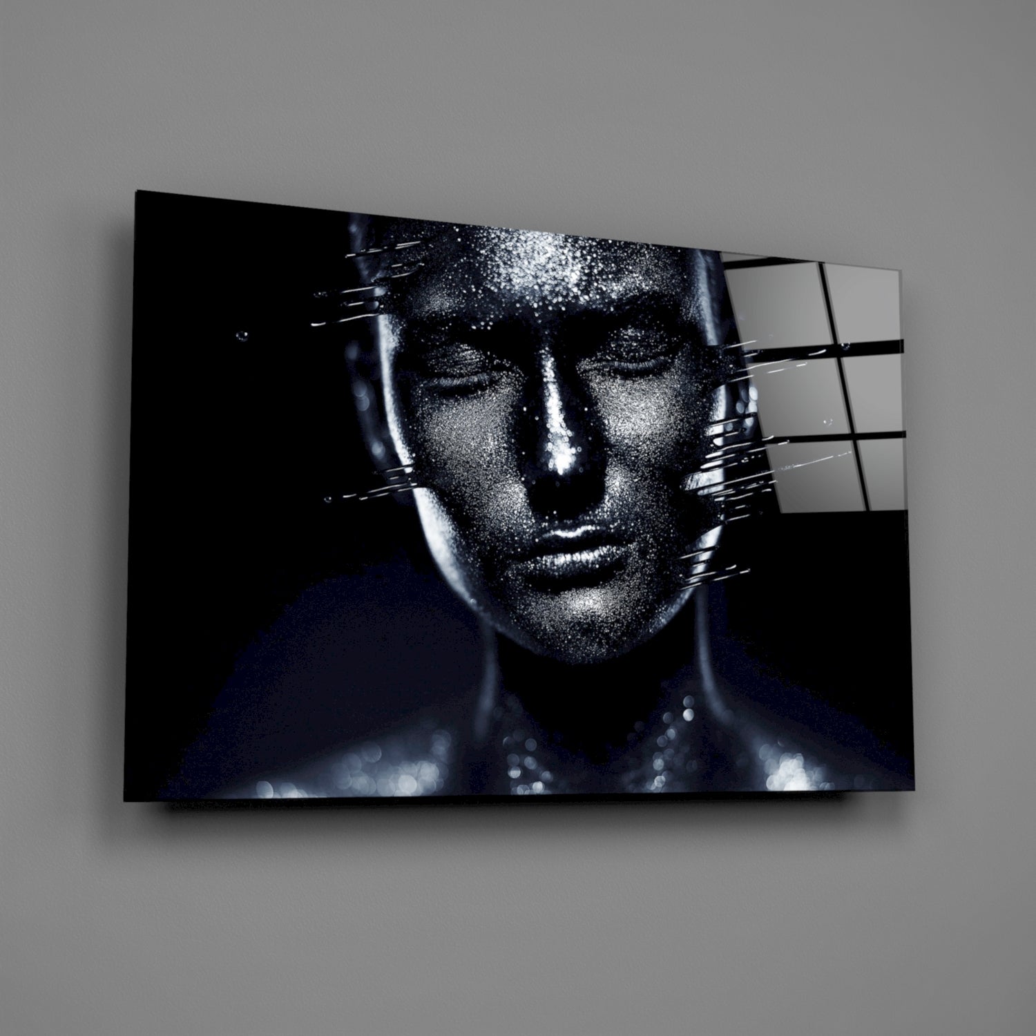 Angled display of glass wall art showing monochromatic female portrait with shimmering texture effect and black grid overlay on gray wall.