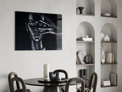 Black and white portrait glass art displaying woman's face with hand across eyes, hung above round black dining table with beige chairs and white shelving unit.