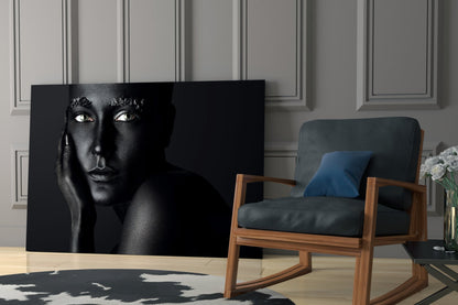 Black glass wall art featuring a woman's portrait with striking white eyes against dark skin, displayed in a modern interior setting beside a wooden chair.