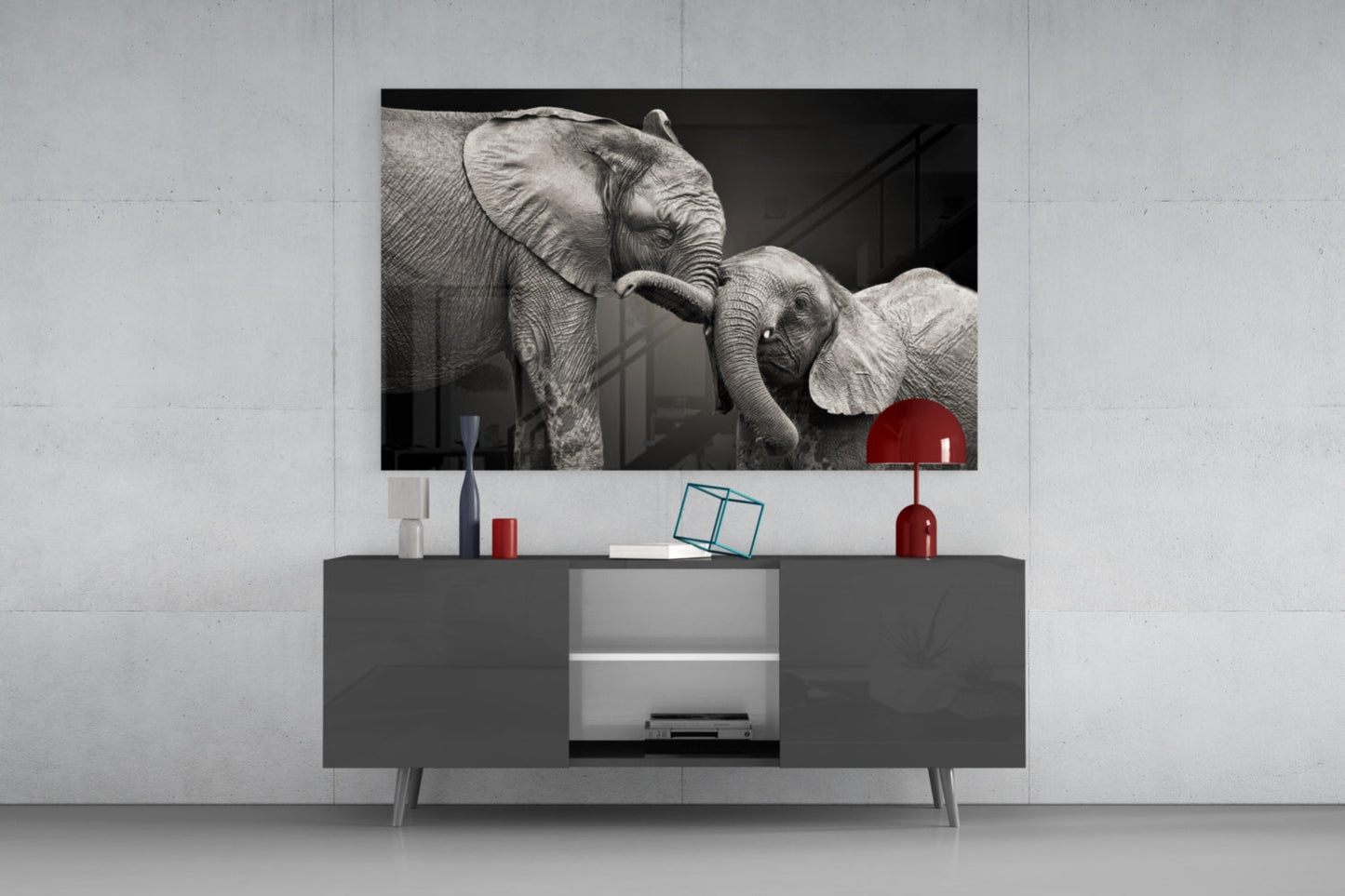 Black and white glass wall art depicting two elephants with intertwined trunks, displayed above a gray media console in a modern interior setting.