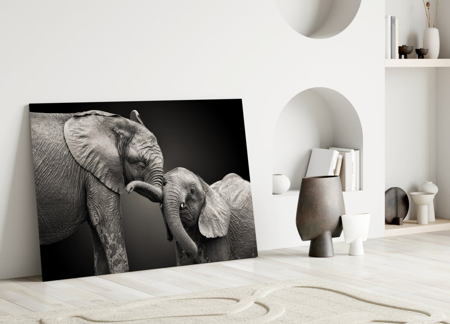 Black and white glass wall art depicting an adult elephant with its trunk touching a baby elephant, displayed leaning against a white interior wall.