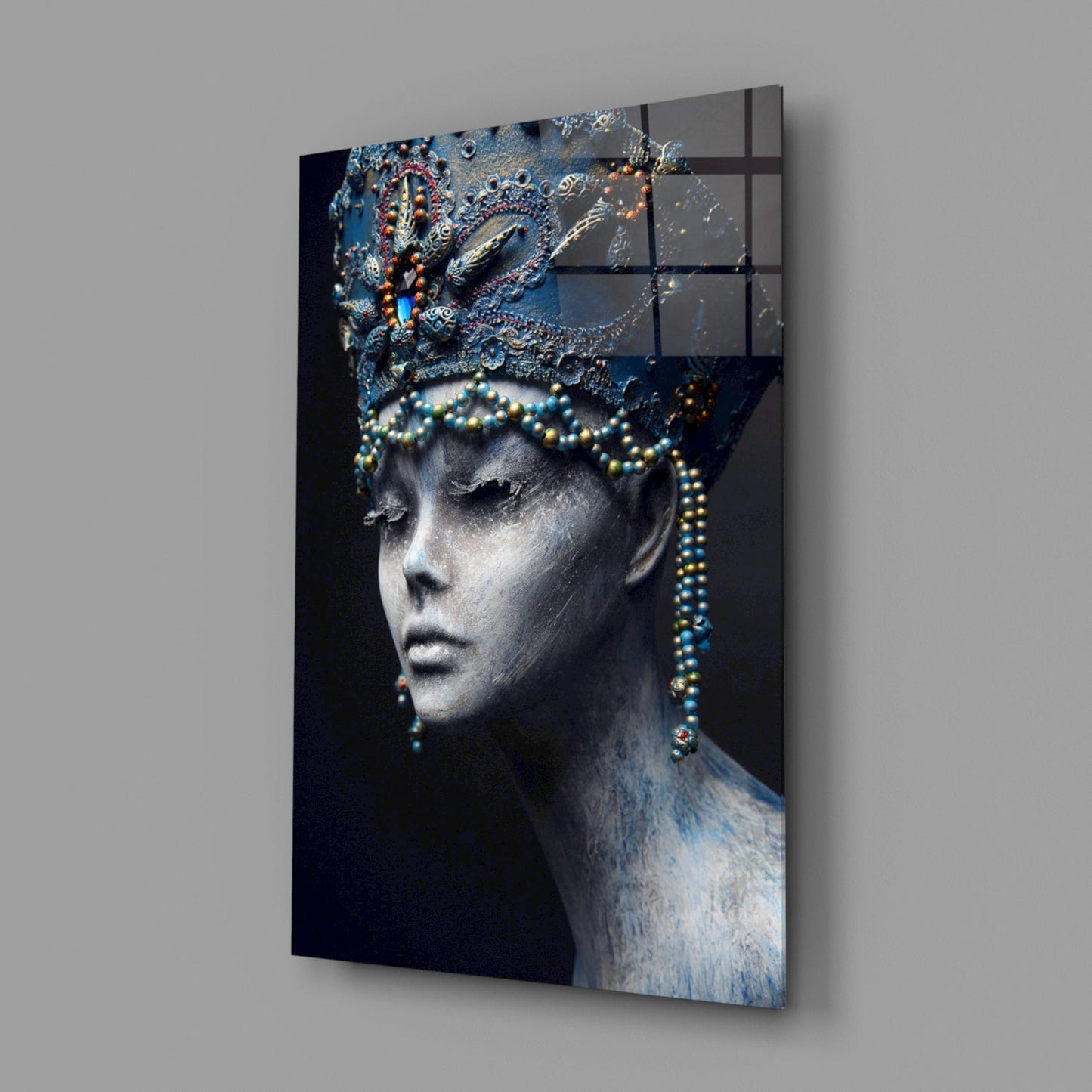 Angled view of glass wall art depicting a gray sculptural female profile wearing an ornate blue headdress and beaded necklaces against a dark background.