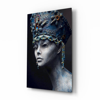 Profile view of a pale face wearing an ornate blue headpiece and beaded necklace against dark background on glass panel.