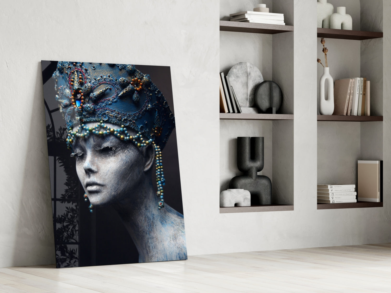 Glass wall art leaning against white wall displays portrait of woman with blue ornate headdress and beaded necklace beside built-in shelving with decorative objects and books.
