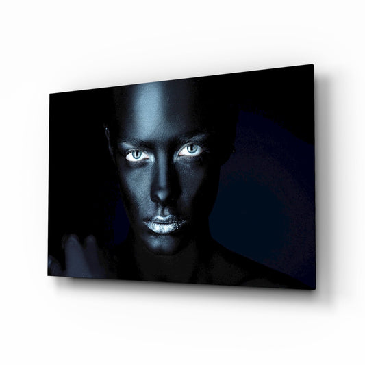 Woman's face with striking blue eyes against dark background on glass wall art, displayed at angle showing depth and shadow.
